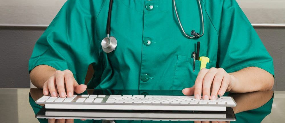 "Operatore sanitario con camice verde e stetoscopio al collo che digita su una tastiera di computer su una scrivania riflettente